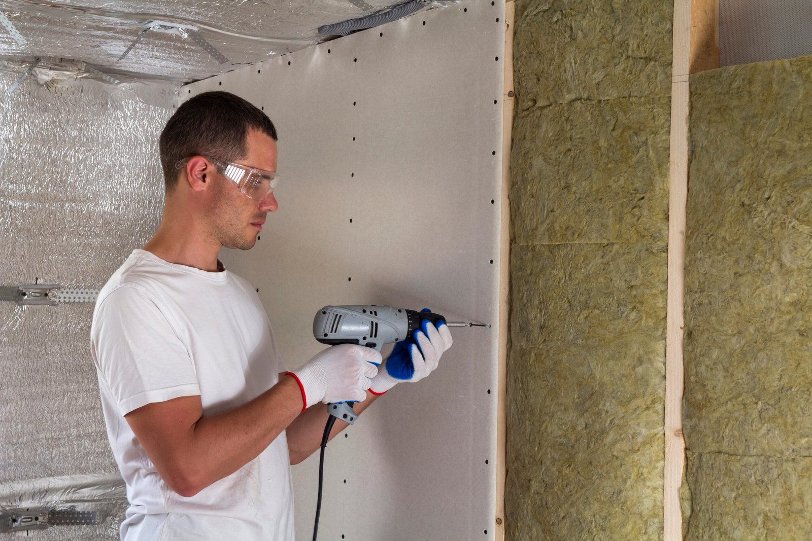Worker in goggles with screwdriver working on insulation. Drywall on wall beams, insulating rock wool staff in wooden frame. Comfortable warm home, economy, construction and renovation concept.